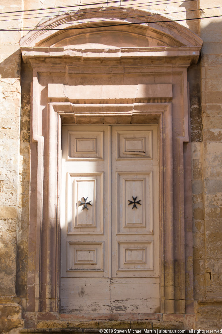 Weathered pale double door with small dark eight-pointed crosses set within a deep projecting limestone surround.