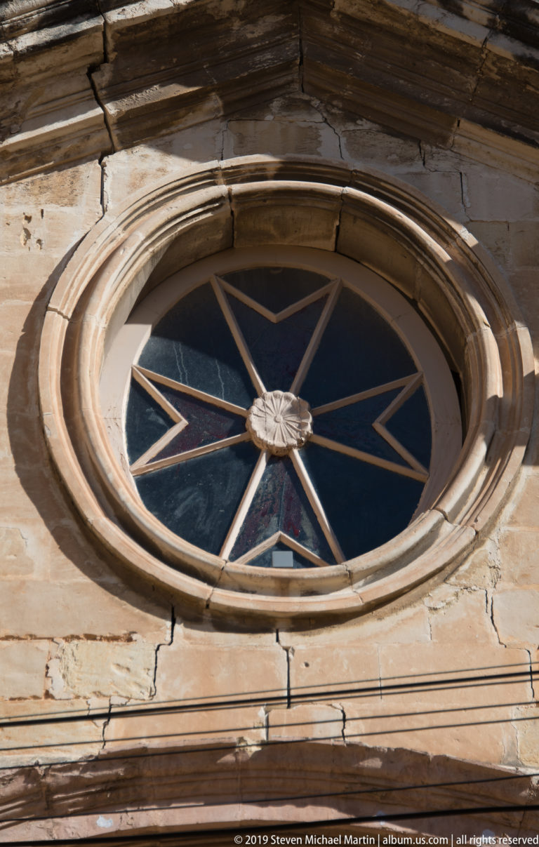 Circular dark-glazed window with pale radial spokes and central boss set within concentric moldings in weathered limestone.