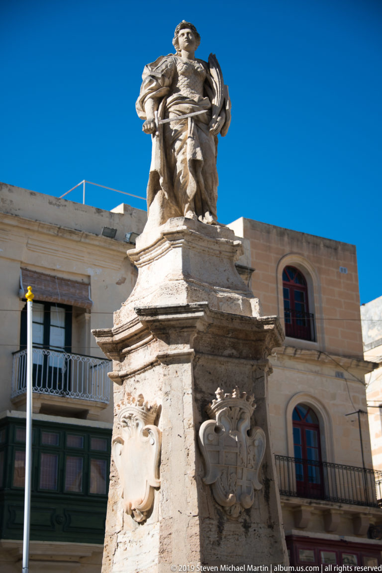 Armored stone figure standing on a tall heraldic pedestal against blue sky, with historic balconies and windows behind.