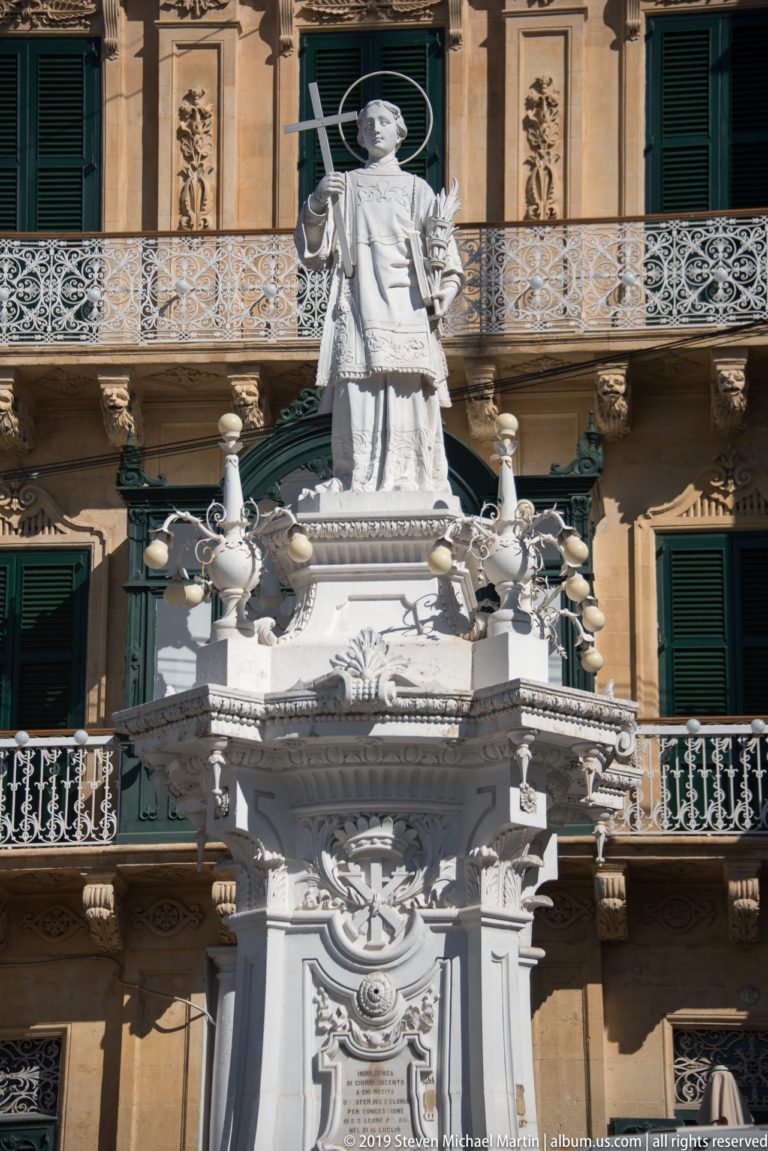 Standing stone figure with long staff atop an ornate white monument, backed by balconies and green shutters.