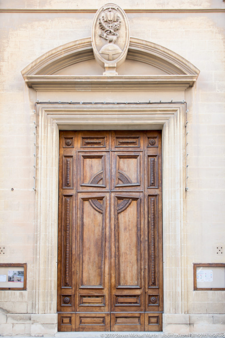 Large paneled wooden double door in a pale limestone surround beneath a triangular pediment and circular carved emblem.