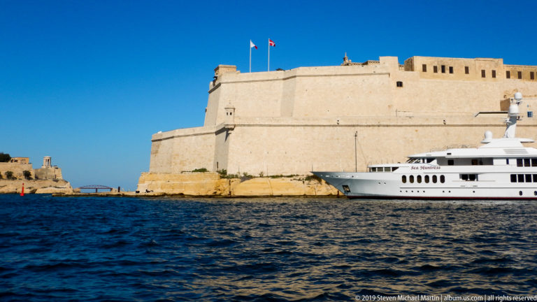 Fort St Angelo's massive limestone walls behind a large white motor yacht on blue Grand Harbour water.