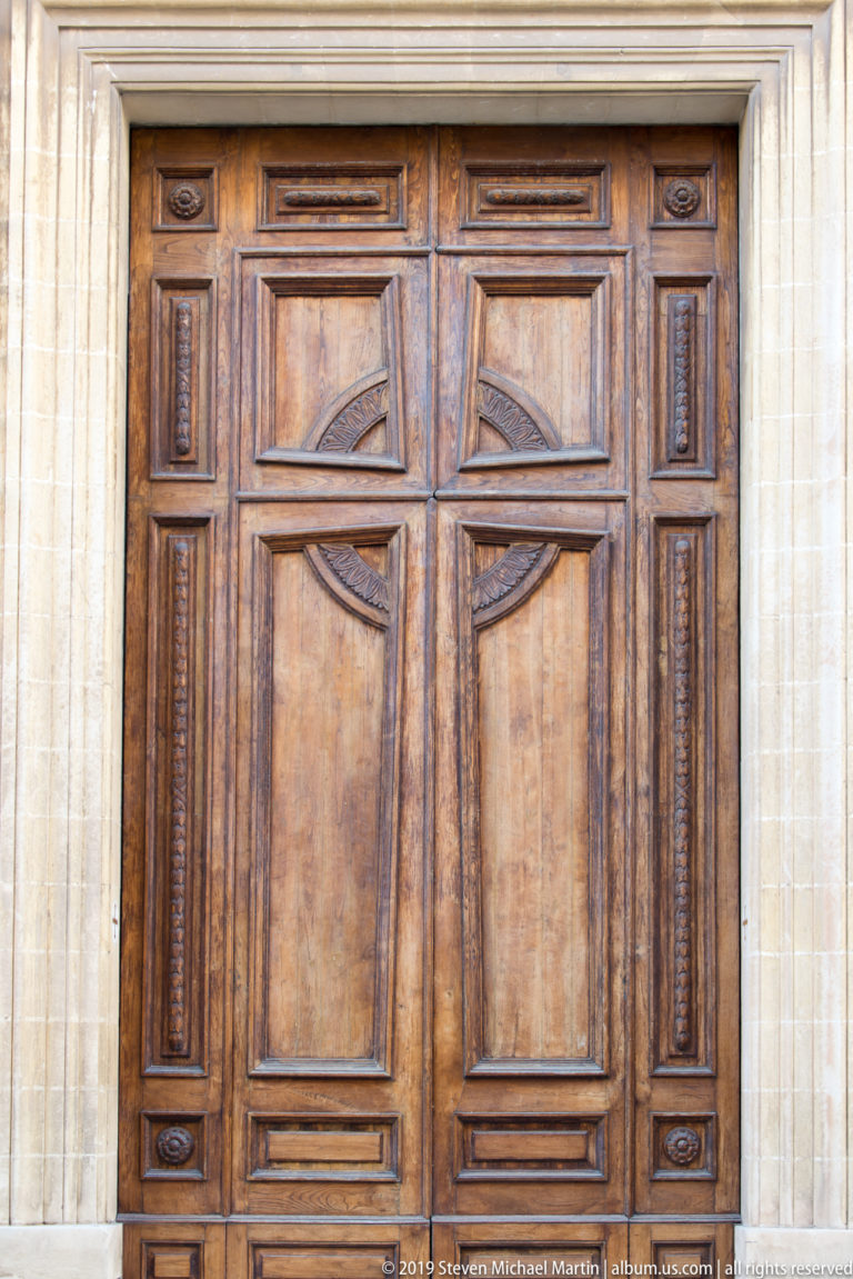 Large paneled wooden double door with curved carved motifs, narrow ornamental side panels, and a pale stone surround.