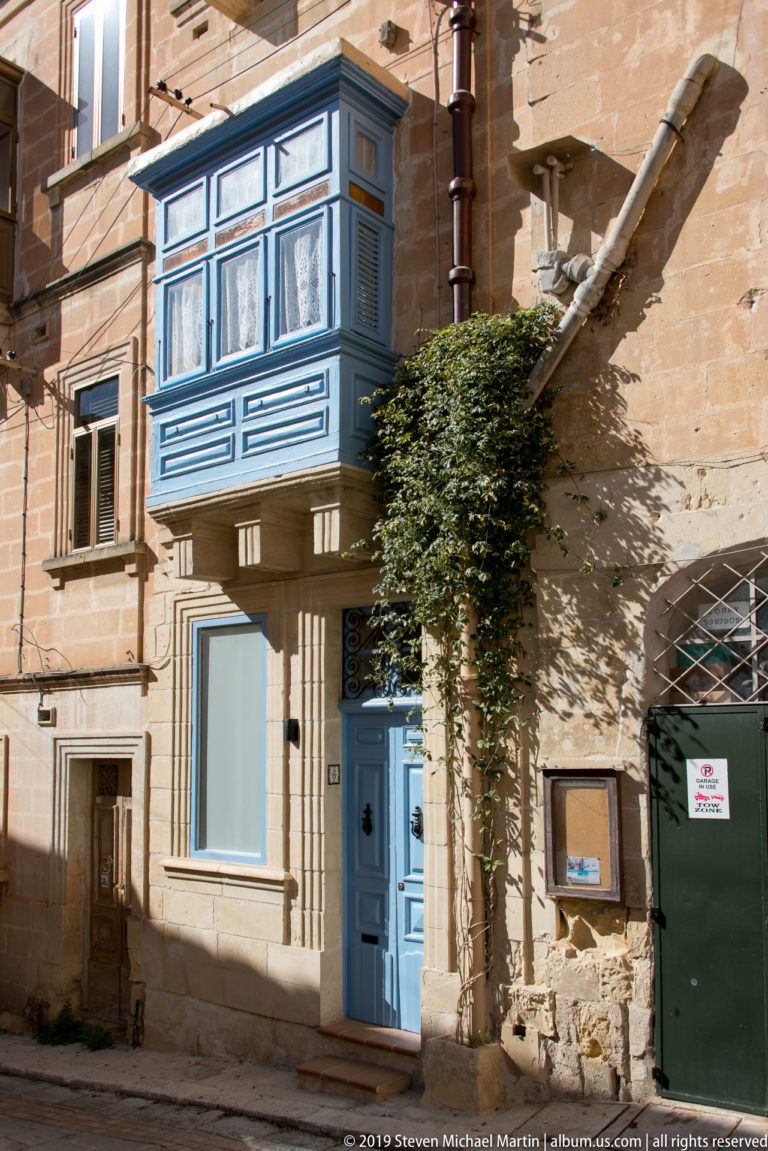 Blue enclosed wooden balcony above a matching blue door on a honey-colored limestone façade with climbing greenery.
