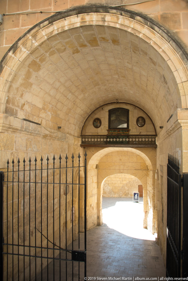 Limestone barrel-vaulted passage with black iron gates and repeated arches leading toward bright daylight at the far end.