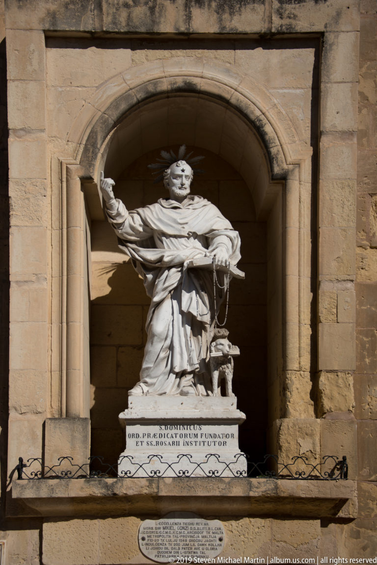 White stone statue of a robed saint with raised arm standing in a deep arched niche above inscribed tablets.