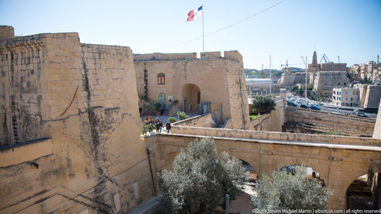 Elevated view of massive limestone fortifications, arched bridge and gateway, Maltese flag, trees, and harbor infrastructure beyond.