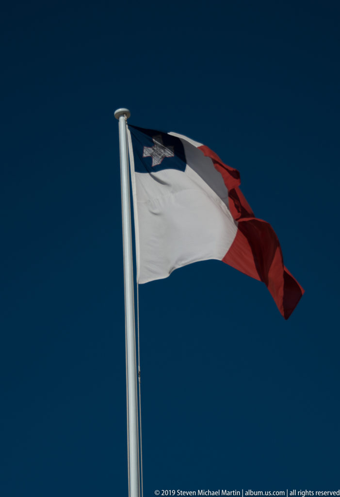 Maltese national flag flying against a deep blue sky, with white and red fields and George Cross at upper hoist.