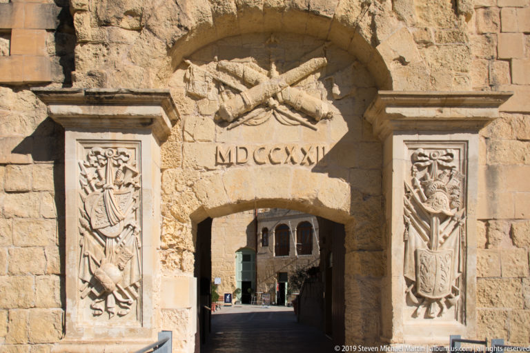 Close view of an arched limestone gateway dated MDCCXXI, with crossed weapons above and carved military trophy panels on both sides.