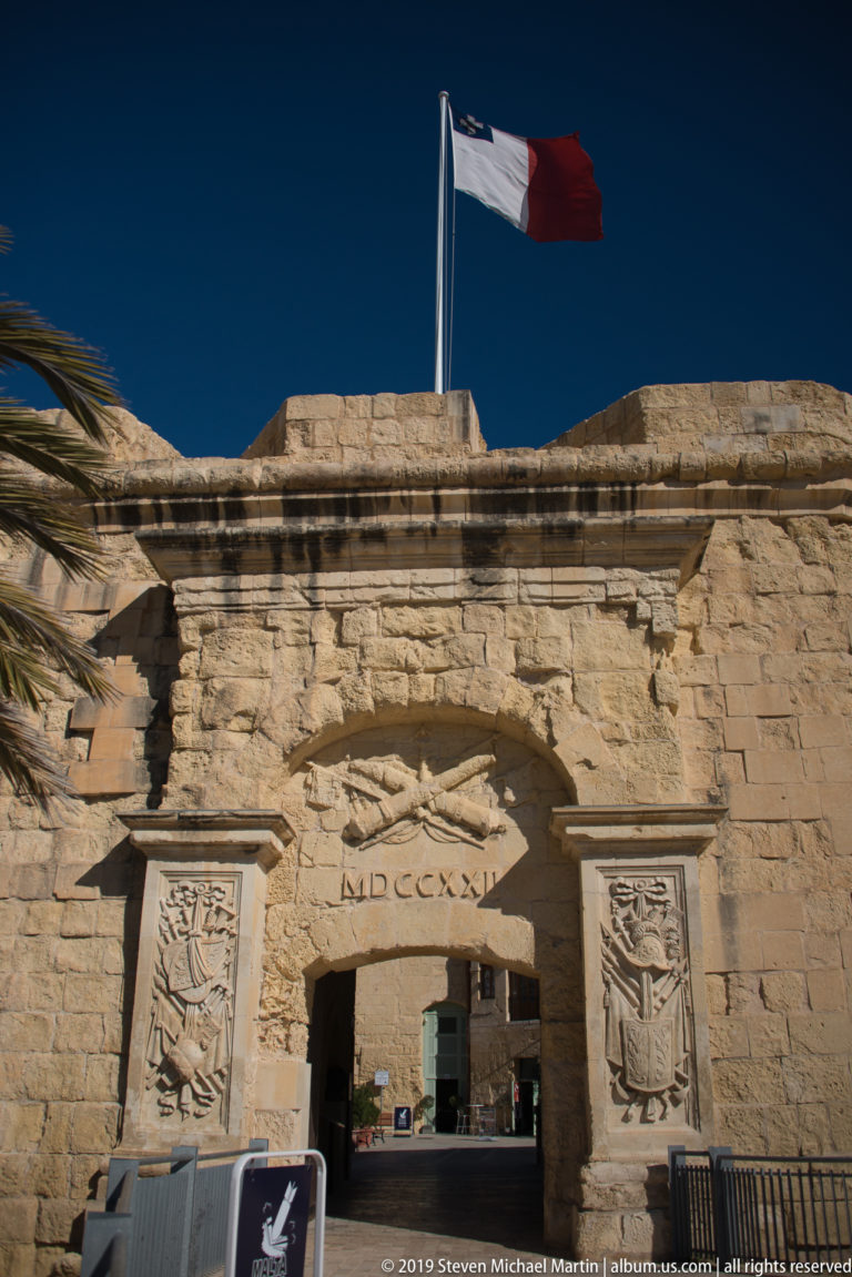 Maltese flag above a rusticated limestone gateway with carved military panels and the Roman date MDCCXXI over the arch.