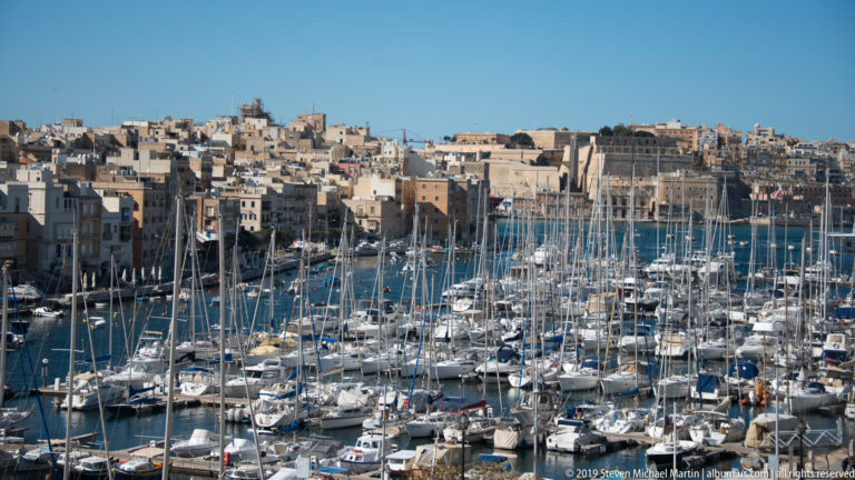 Dense marina filled with white yachts and tall masts below a hillside of tightly packed limestone buildings.