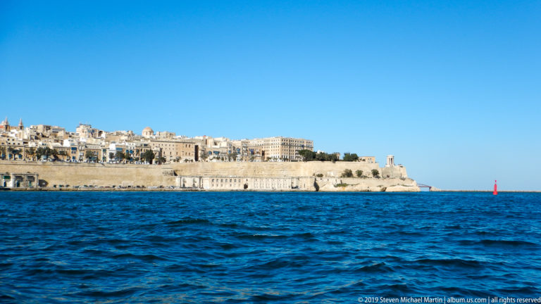 Fortified limestone peninsula of Valletta above deep blue harbor water, with dense buildings and a red navigation marker at right.