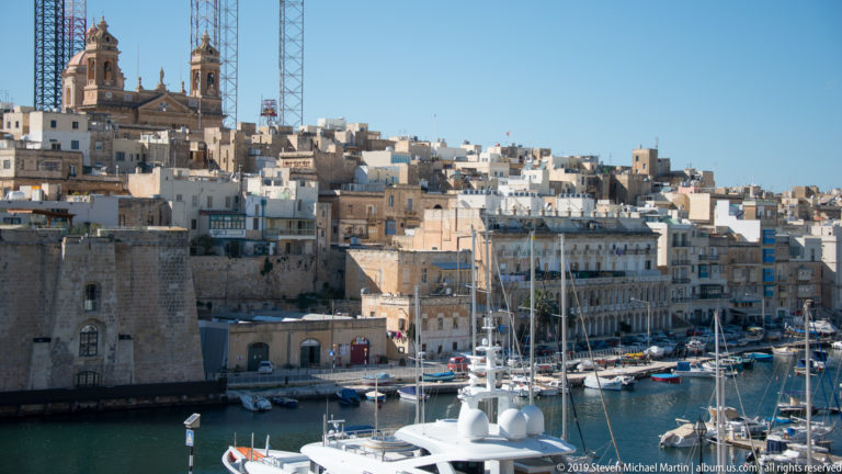 Elevated view over marina yachts toward dense honey-colored limestone buildings and a massive fortified wall at left.