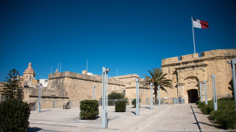 Maltese flag above a massive limestone fort entrance with arched gateway, paved forecourt, palm tree, and modern posts.