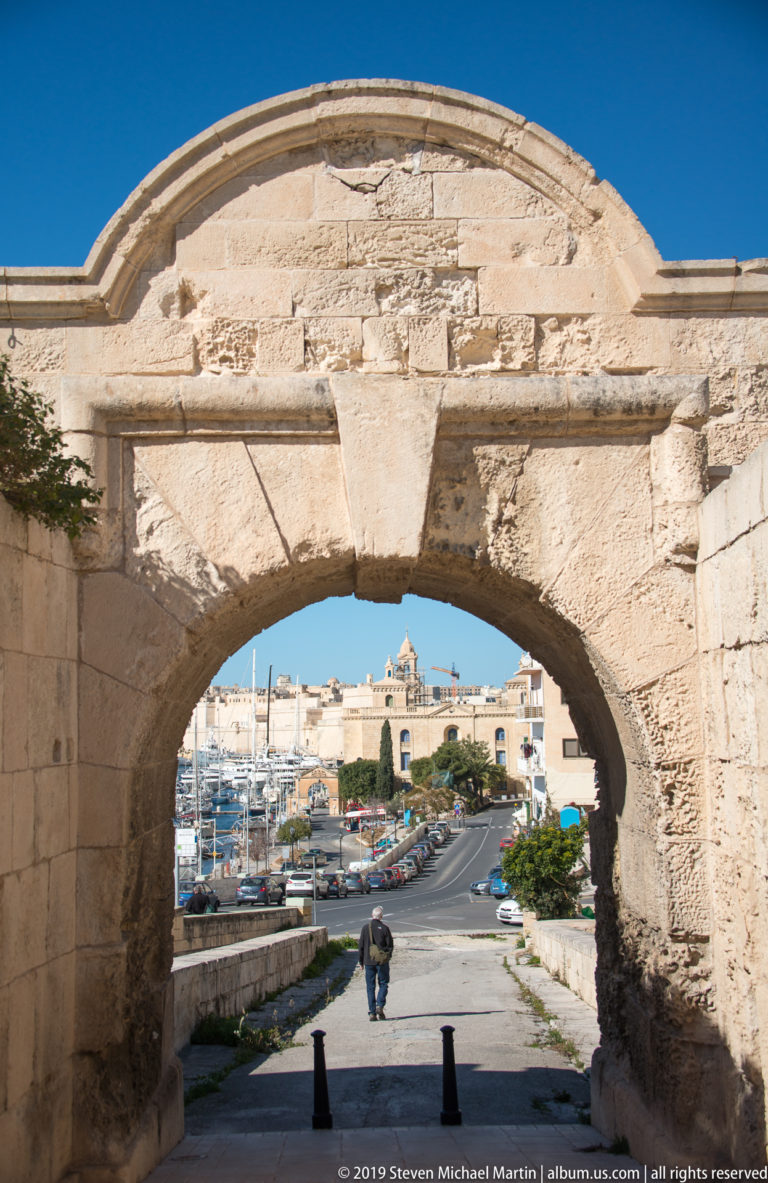 Large weathered limestone arch framing a road, parked cars, marina masts, and harbor-side buildings beyond.