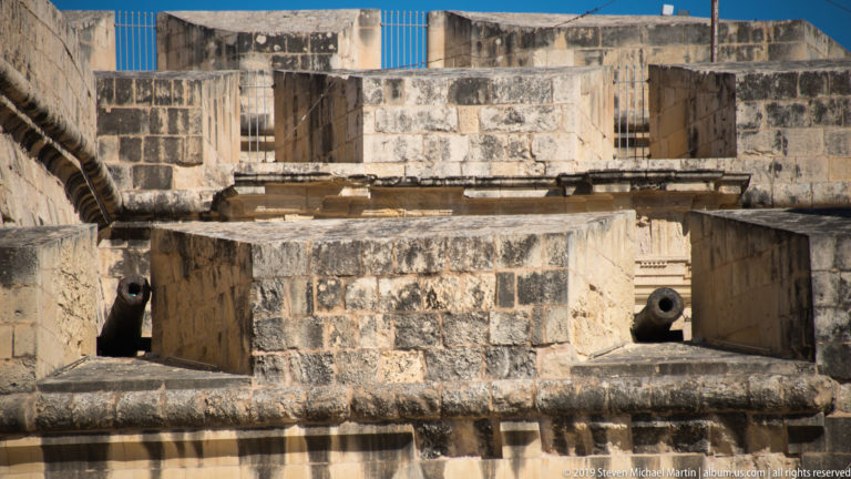 Two cannon barrels projecting from rectangular embrasures in thick, weathered limestone fortifications with stepped masonry above.