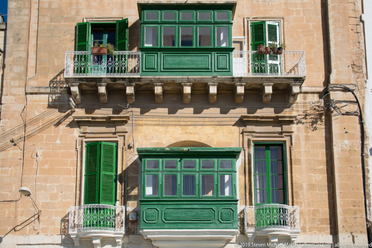 Two green enclosed wooden balconies with matching shutters and metal railings on a honey-colored limestone façade.