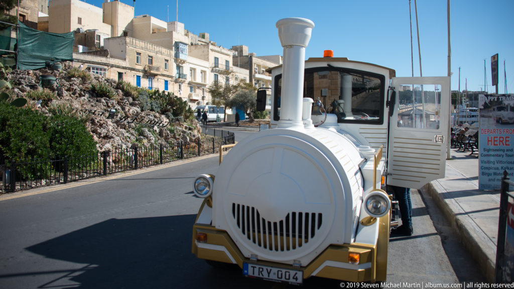White locomotive-shaped tourist road train parked beside a limestone streetscape with rocky landscaping and harbor development nearby.