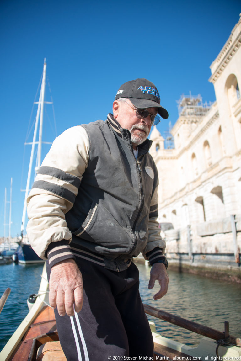 Boatman in cap and jacket holding a line aboard a wooden craft, with yachts and limestone waterfront buildings behind.