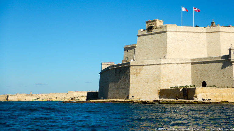 Massive pale limestone walls of Fort St Angelo seen from water level, with flags flying above the upper fortress.