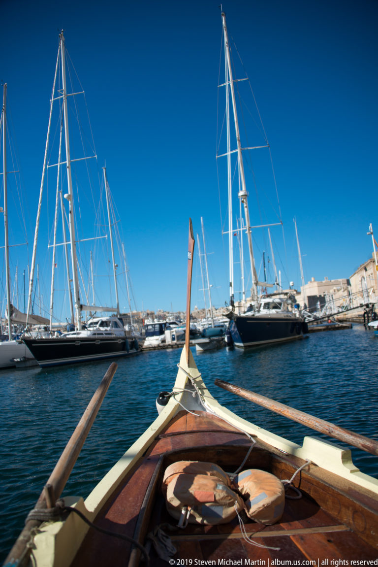 Pointed wooden boat bow with long oars aimed toward a marina filled with sailboats and tall masts under blue sky.