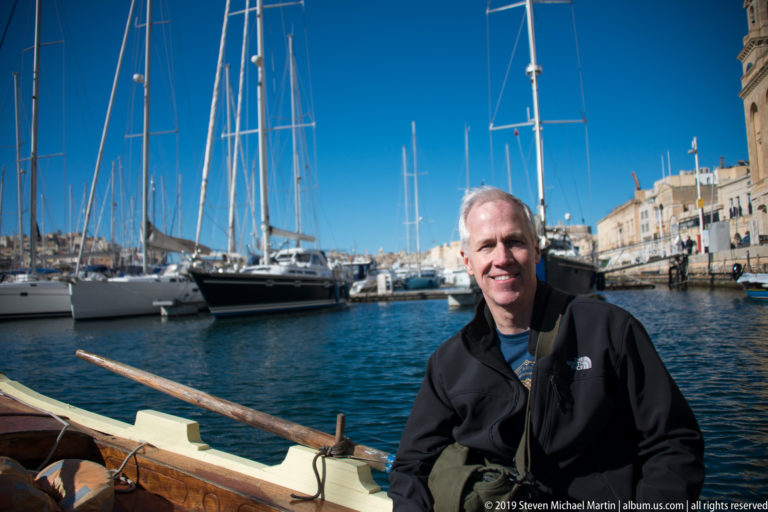 Smiling man seated in a wooden harbor boat with an oar, sailboats, marina water, and limestone buildings behind.