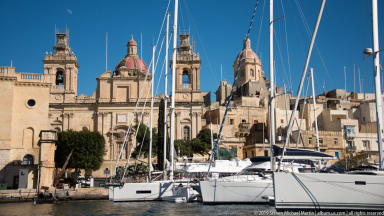 Ornate limestone church with twin bell towers and red domes behind white yachts and a dense screen of masts.