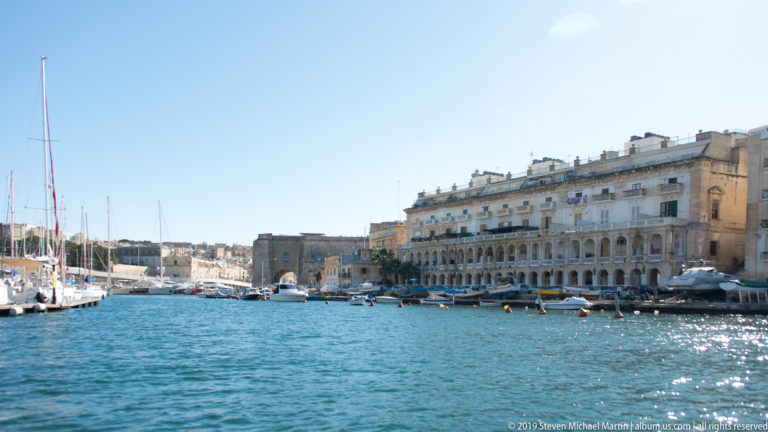 Long limestone waterfront building with repeated arches and balconies beside blue harbor water, yachts, and a marina.