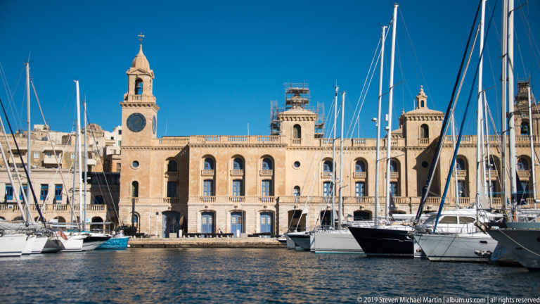 Long limestone Malta Maritime Museum building with central clock tower behind yachts and tall masts in Vittoriosa marina.