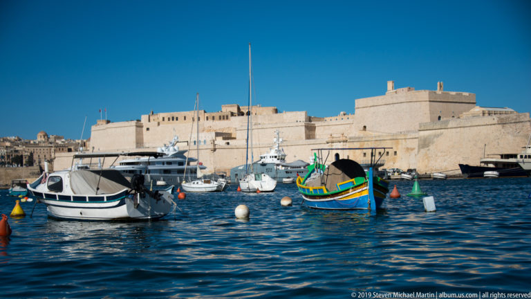 Colorful traditional Maltese boat among white pleasure craft, with the massive limestone walls of Fort St Angelo behind.