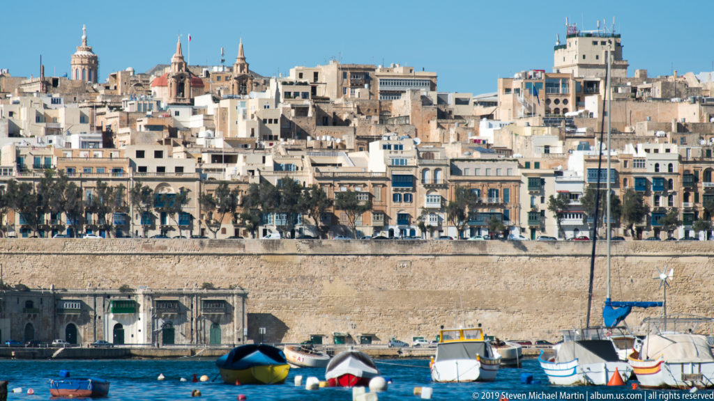 Small boats in a marina below Vittoriosa's long limestone fortification wall, dense hillside buildings, balconies, and church towers.