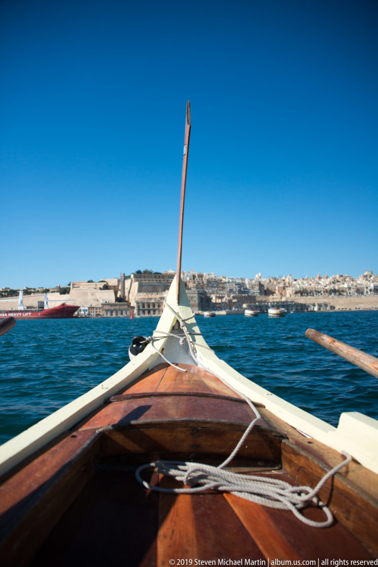 Varnished wooden bow of a traditional Maltese harbor boat with rope and oar, pointing across blue water toward a limestone shoreline.