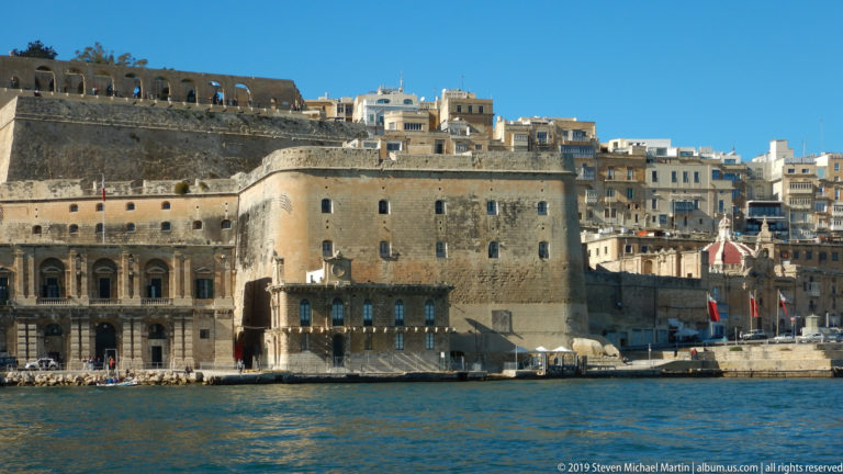 Large limestone fortifications rising from Grand Harbour, with arched openings, flags, waterfront terraces, and dense buildings behind.