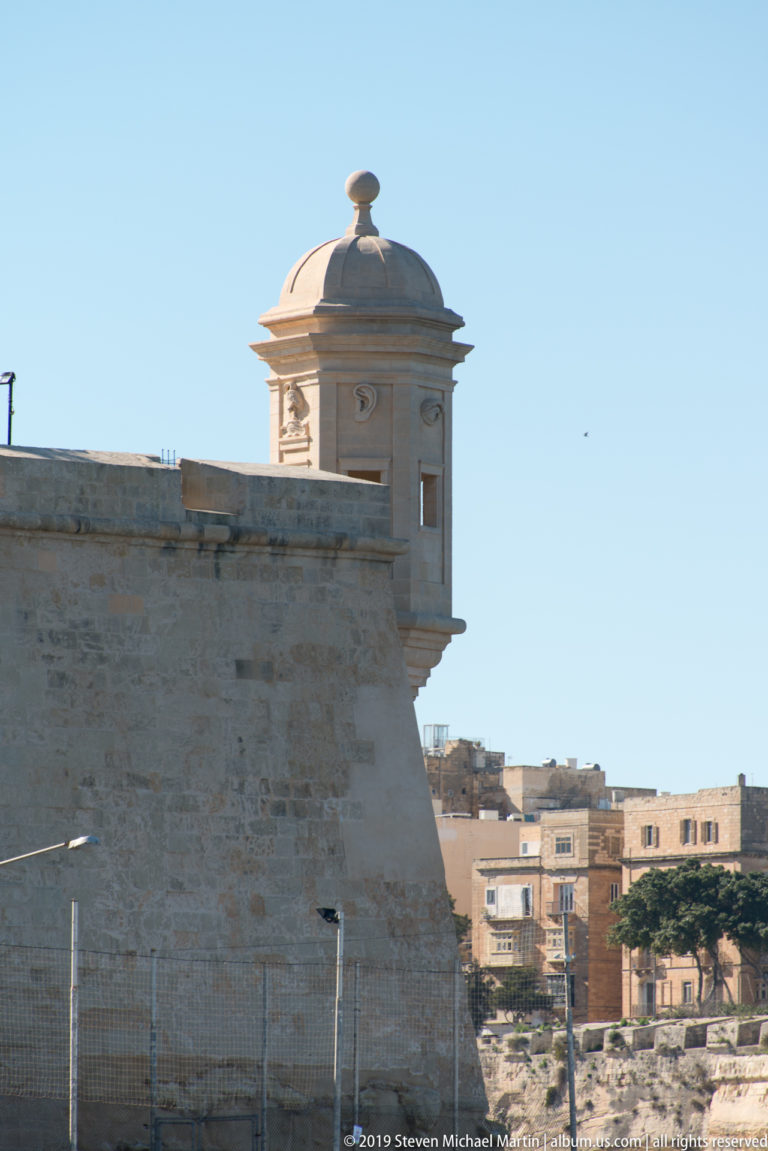 Domed stone sentry box projecting from the corner of a pale limestone fortification wall above the harbor.