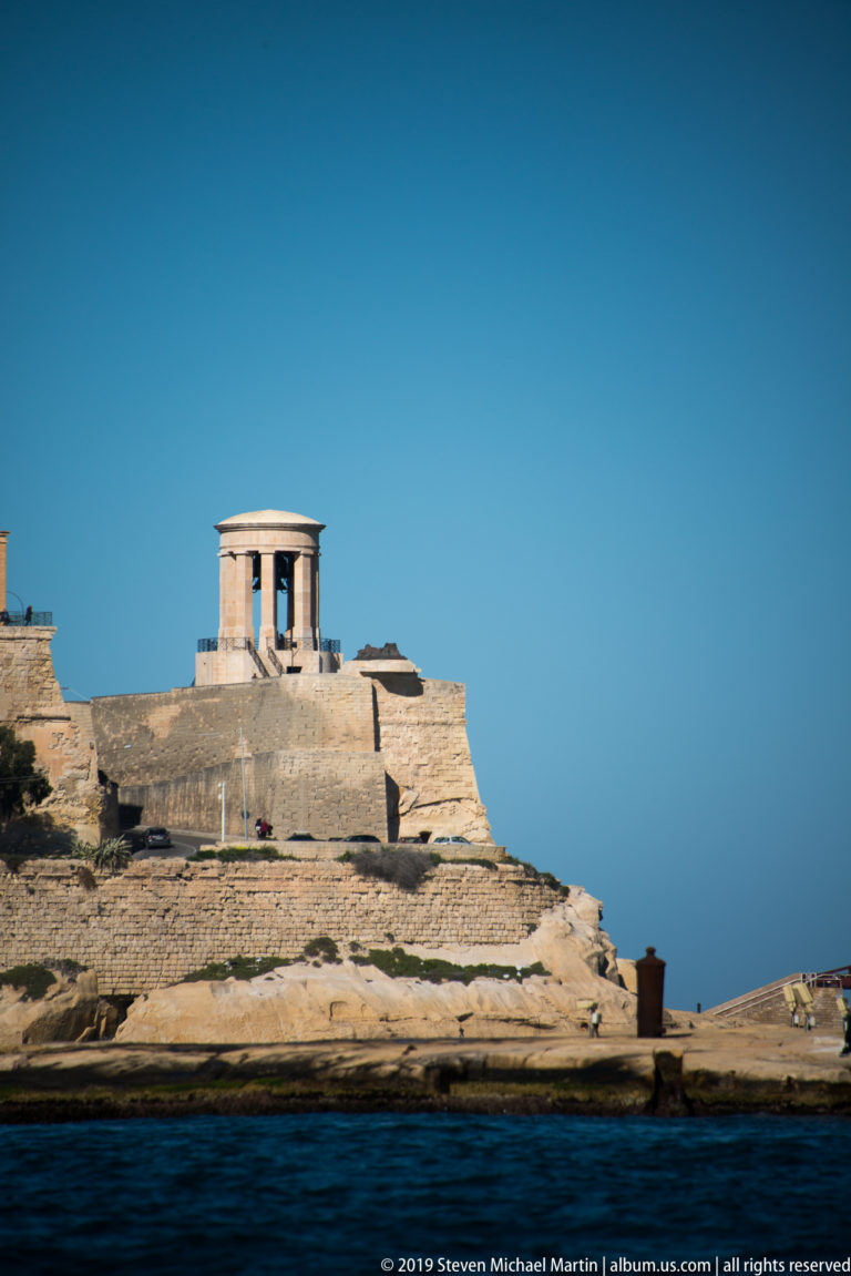 Classical open-sided stone pavilion of the Siege Bell Memorial standing on Valletta's rocky bastions beneath a clear blue sky.