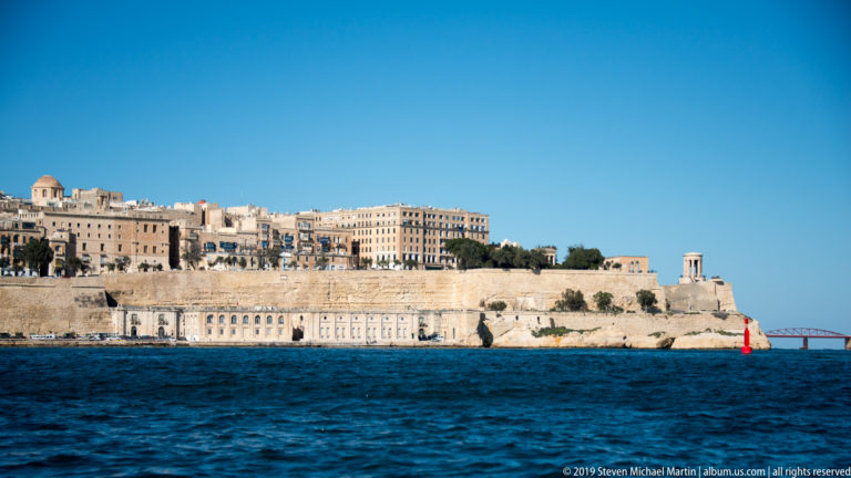 Long limestone fortification wall beneath Valletta buildings, seen from the water with a small classical lookout structure near the bastion edge.