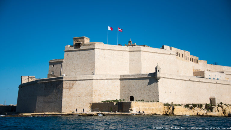 Massive pale limestone bastions of Fort St Angelo rising from the water, with flags flying above the upper fortifications.