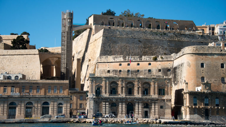 Historic limestone waterfront buildings at Vittoriosa backed by massive fortification walls and upper-city structures.