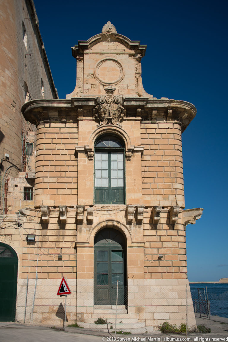 Small ornate limestone building with arched windows, carved cornices, central parapet, and adjoining fortified wall under a blue sky.