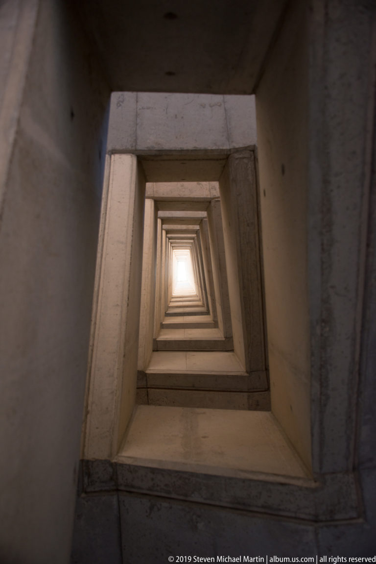 Nested rectangular limestone openings recede upward toward a bright square of daylight inside a massive fortified structure.