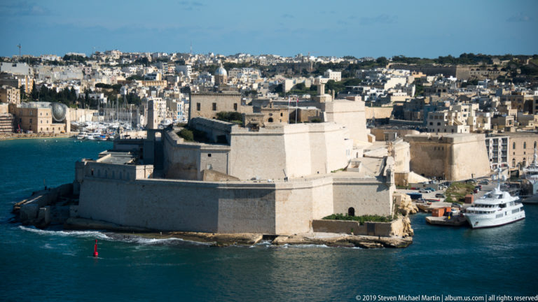 Massive limestone walls of Fort St Angelo project into Grand Harbour, with Vittoriosa buildings and moored vessels behind.