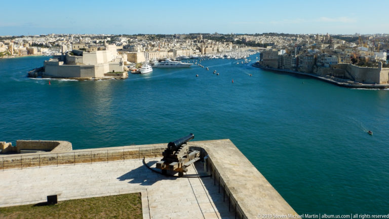 Single historic cannon on a high terrace overlooking Fort St Angelo, blue Grand Harbour, Vittoriosa, and marina basins.