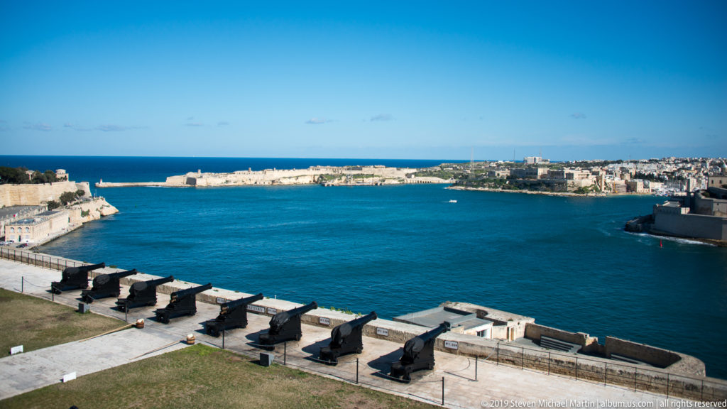 Historic cannon along a Valletta battery overlooking Grand Harbour, low limestone fortifications, and the open Mediterranean horizon.