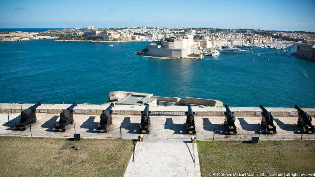 Row of historic black cannon on a stone battery overlooking bright blue Grand Harbour and fortified urban shorelines.