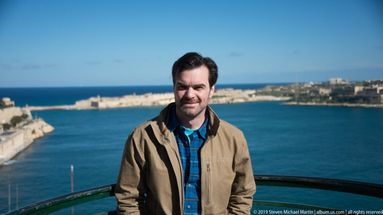 Man in a tan jacket standing at a railing above Grand Harbour, with blue water and limestone fortifications behind.
