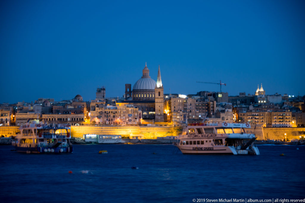 Wide view of Valletta across Marsamxett Harbour, with dome and church spire above illuminated waterfront buildings and ferries.