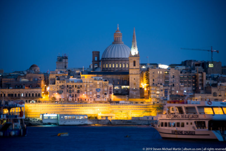 Valletta skyline across blue harbor water, with a large dome, pointed church spire, illuminated bastions, and passenger boat in front.