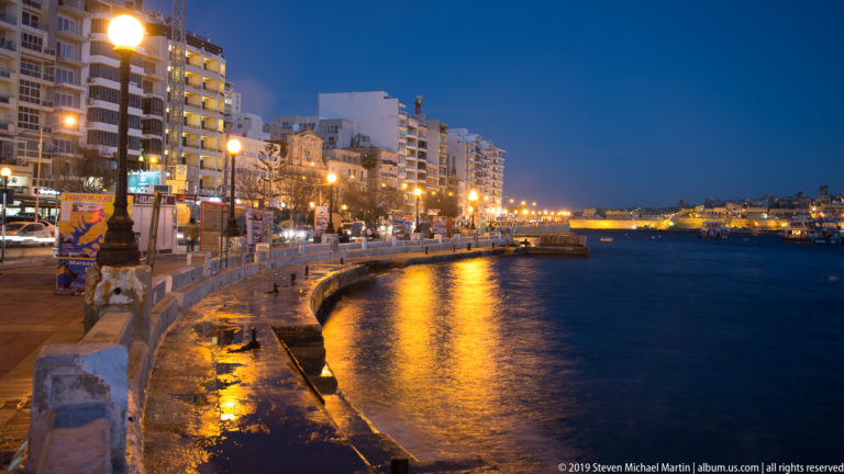 Curving Sliema waterfront at blue hour, with streetlights reflecting on Marsamxett Harbour and apartment buildings lining the promenade.