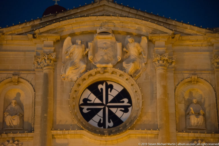 Close view of a limestone church façade with paired sculptures, heraldic shield, circular patterned window, saint niches, and carved capitals.