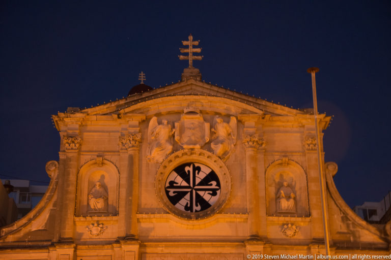 Illuminated limestone church façade with triangular pediment, cross, sculpted figures, saint niches, and a circular black-and-white window.
